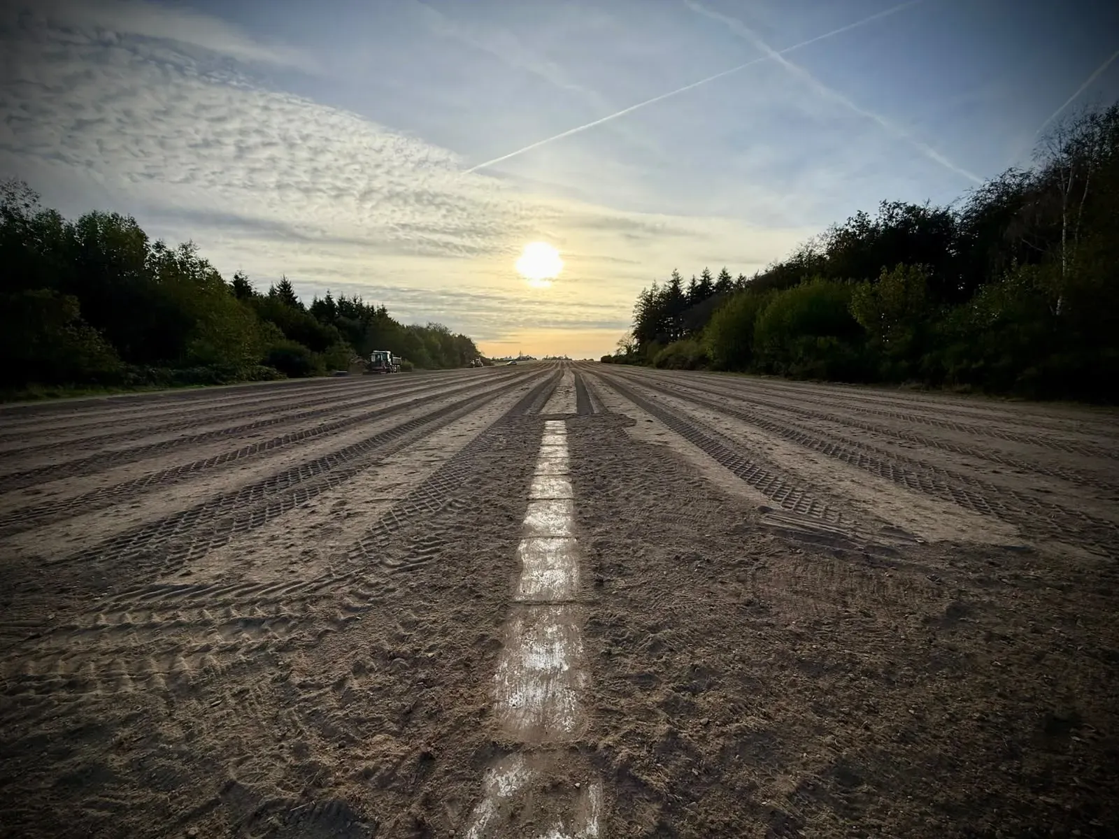 The bare runway at sunset, seen along a painted centreline marker, with machines at the far end between the trees