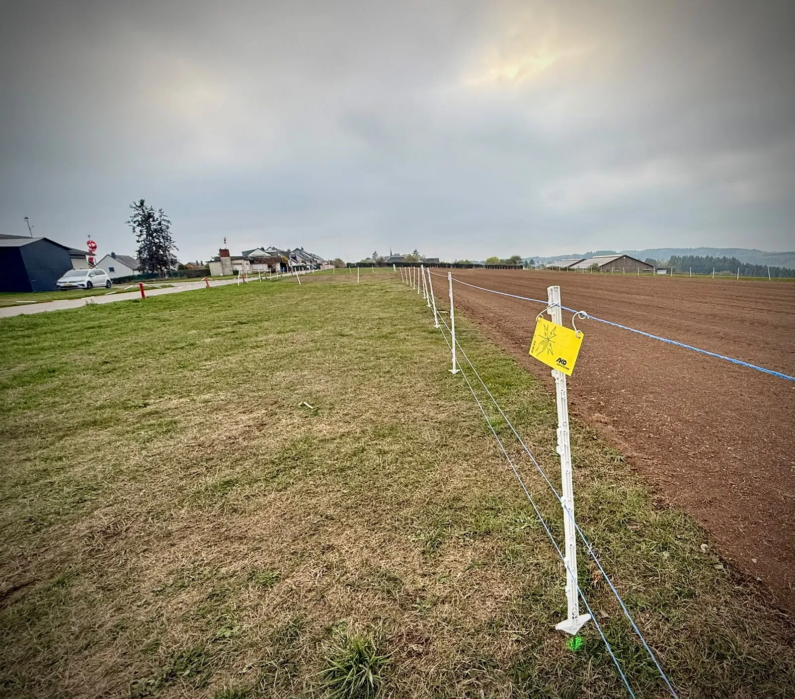 Electric fence posts and strands with a yellow warning sign beside the runway, with grass on the left and prepared soil on the right