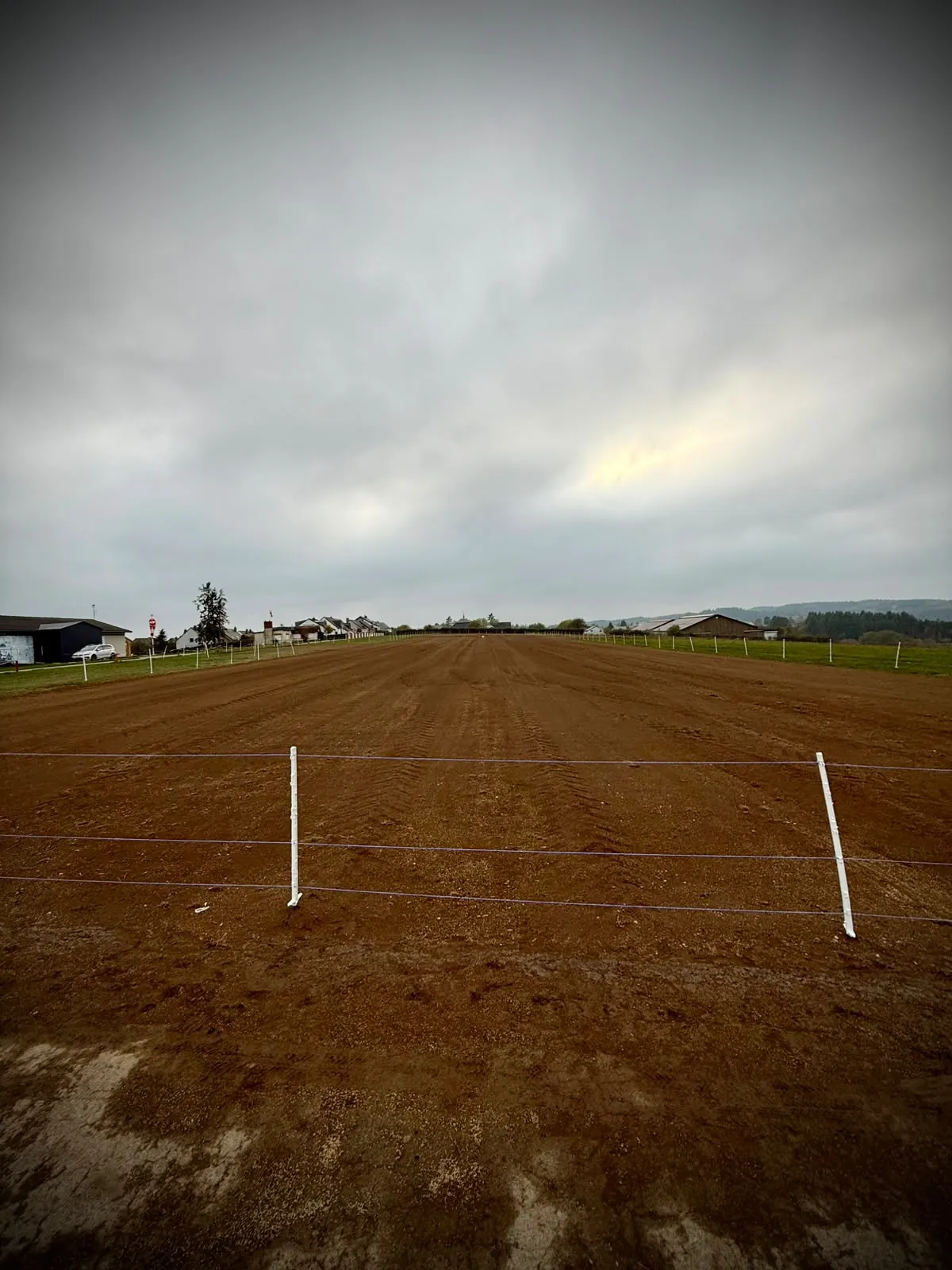 The bare-earth runway behind white electric fence posts at the near end, with hangars in the distance under an overcast sky
