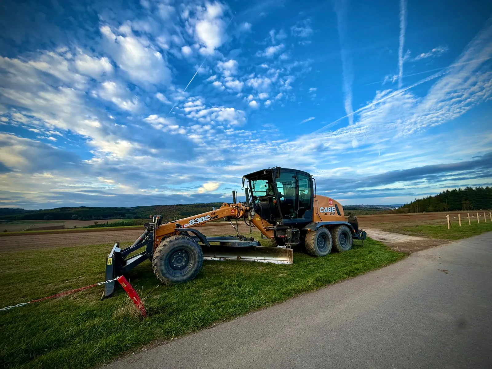 A yellow grader parked on the grass beside the runway under a blue sky with clouds