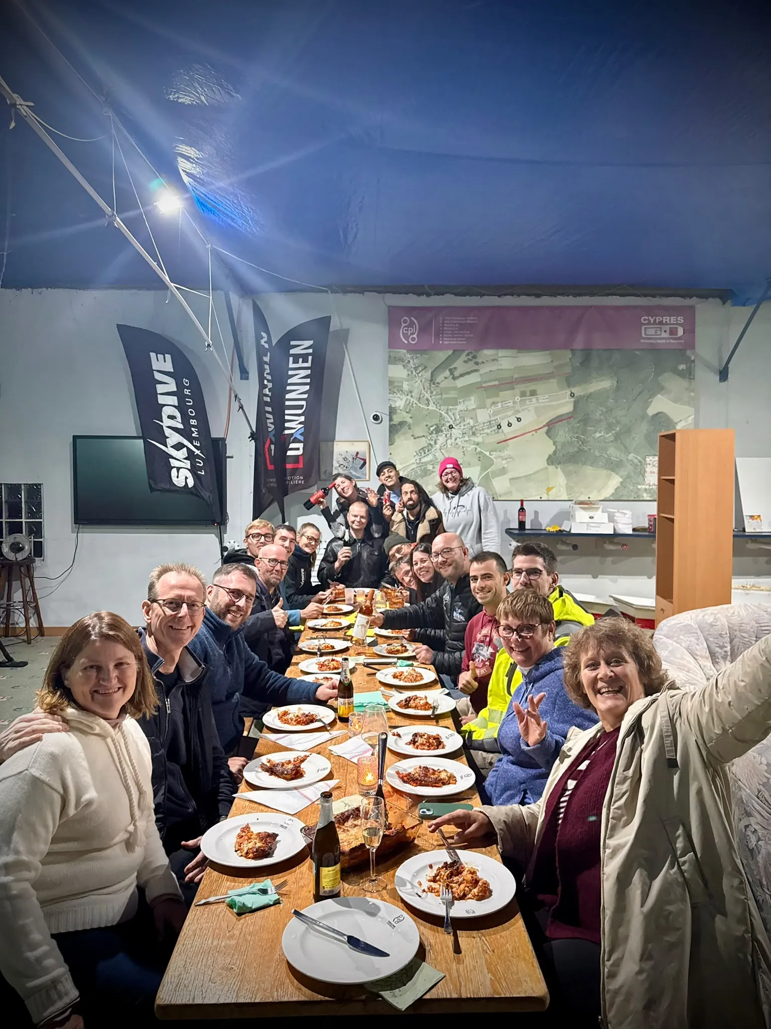 A group smiling towards the camera around a long dinner table in the club hangar, with plates, glasses and flags visible